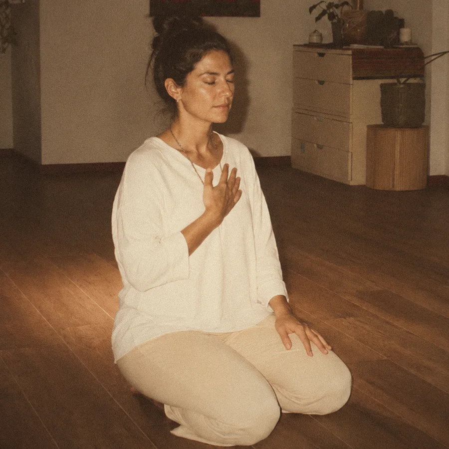 A woman kneels on the studio floor, eyes closed, one hand resting on her heart.